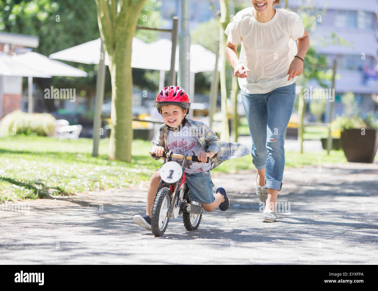 Mother chasing son riding bicycle with helmet in sunny park Stock Photo