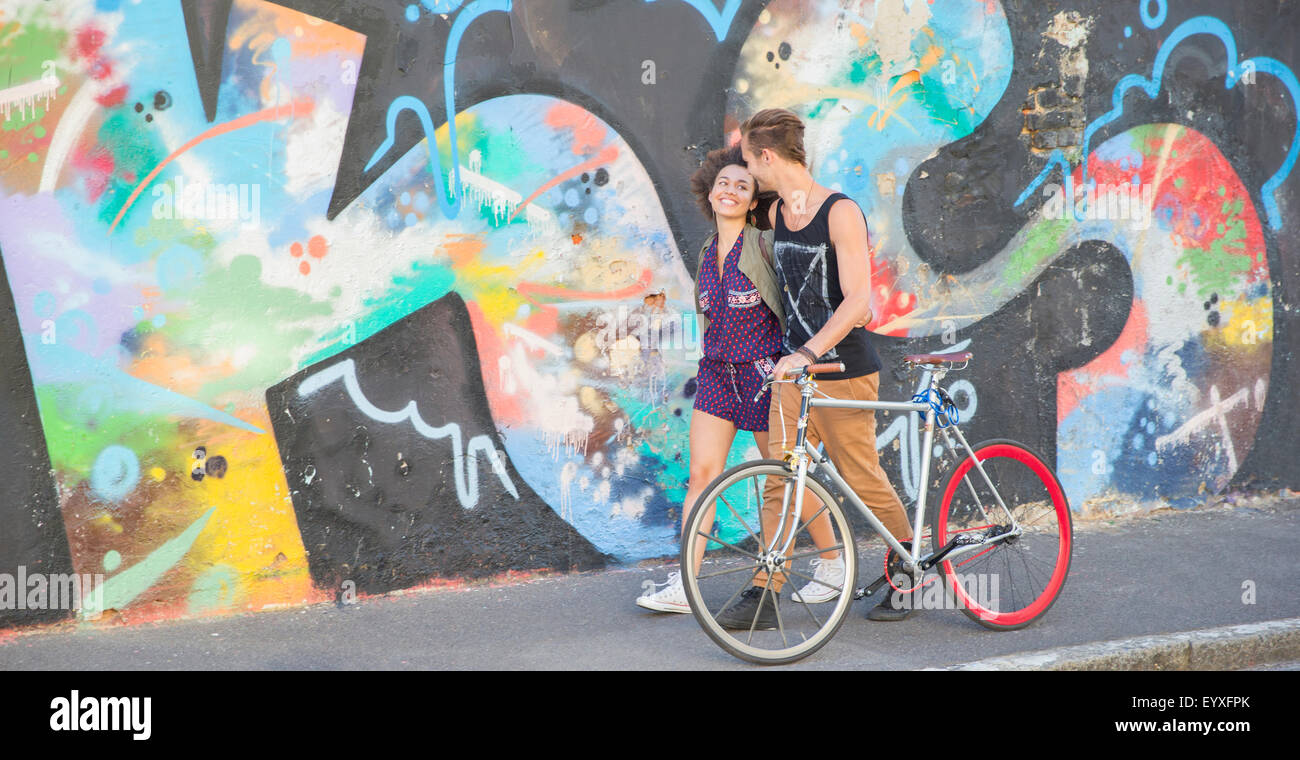 Young couple with bicycle hugging and walking along urban multicolor graffiti wall Stock Photo