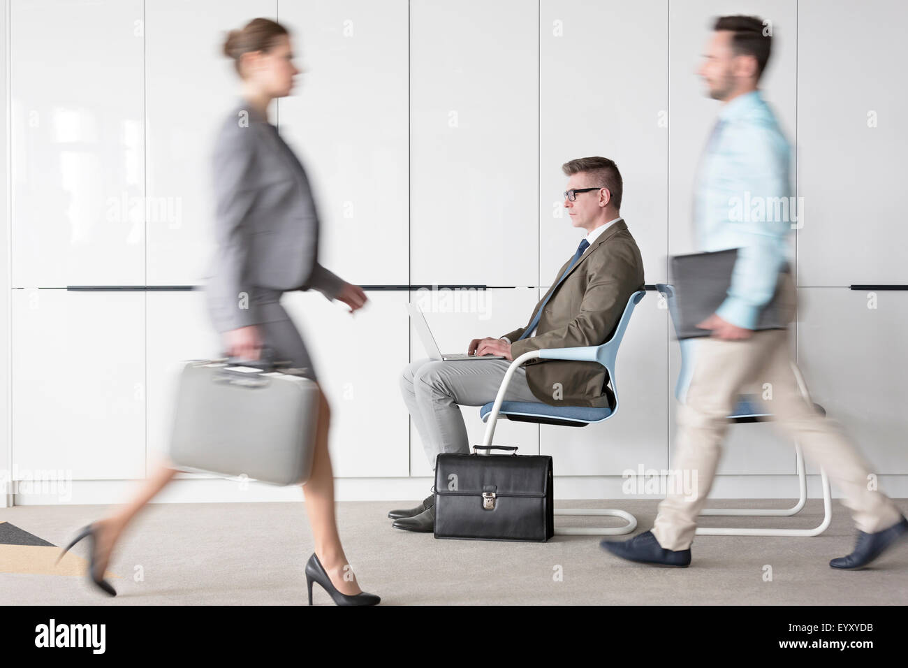 Businessman working at laptop in lobby behind business people on the move Stock Photo