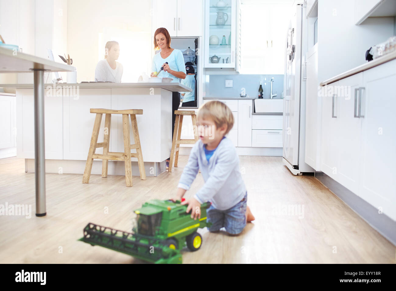 Women cooking in kitchen while boy plays with toy tractor on floor Stock Photo