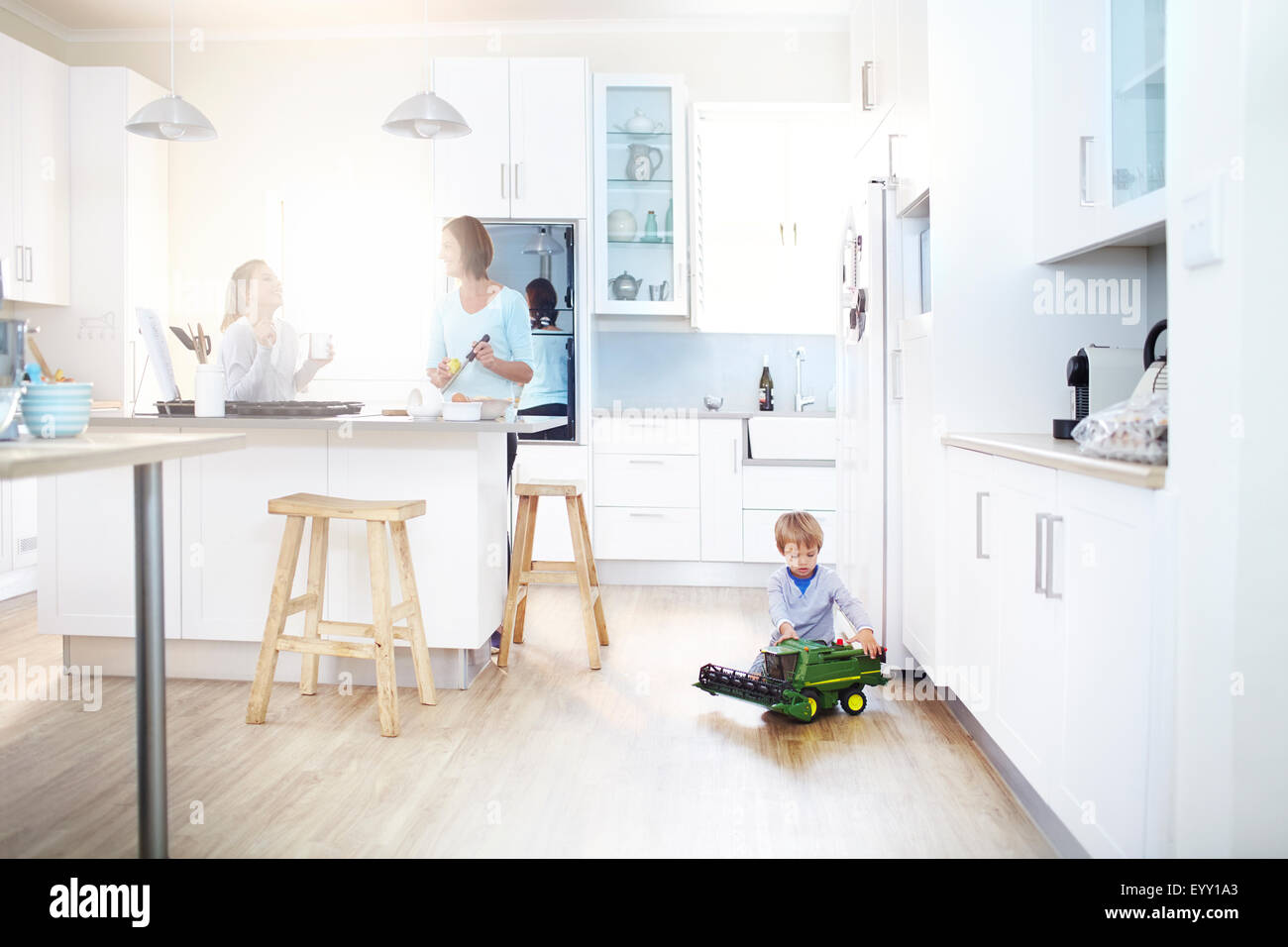 Women cooking in kitchen while boy plays with toy tractor on floor Stock Photo