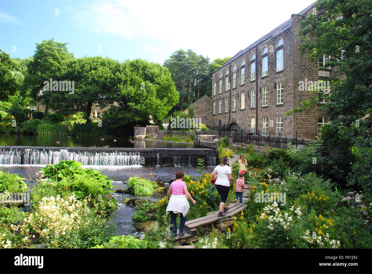 People walk over the wooden public footbridge by Bamford Mill and weir in the Peak District Derbyshire England UK - summer Stock Photo