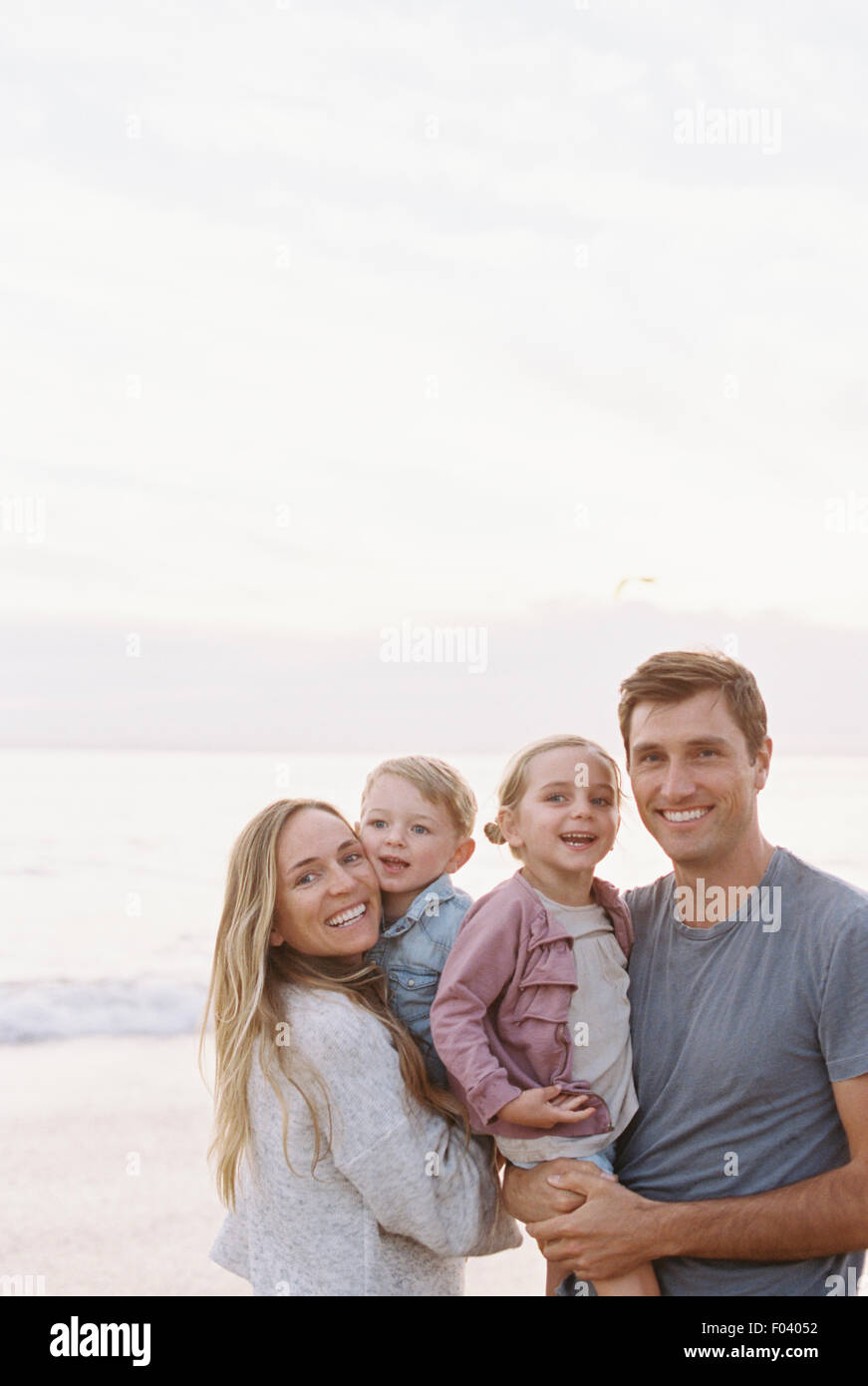 Couple standing with their son and daughter on a sandy beach by the ocean, looking at camera, smiling. Stock Photo
