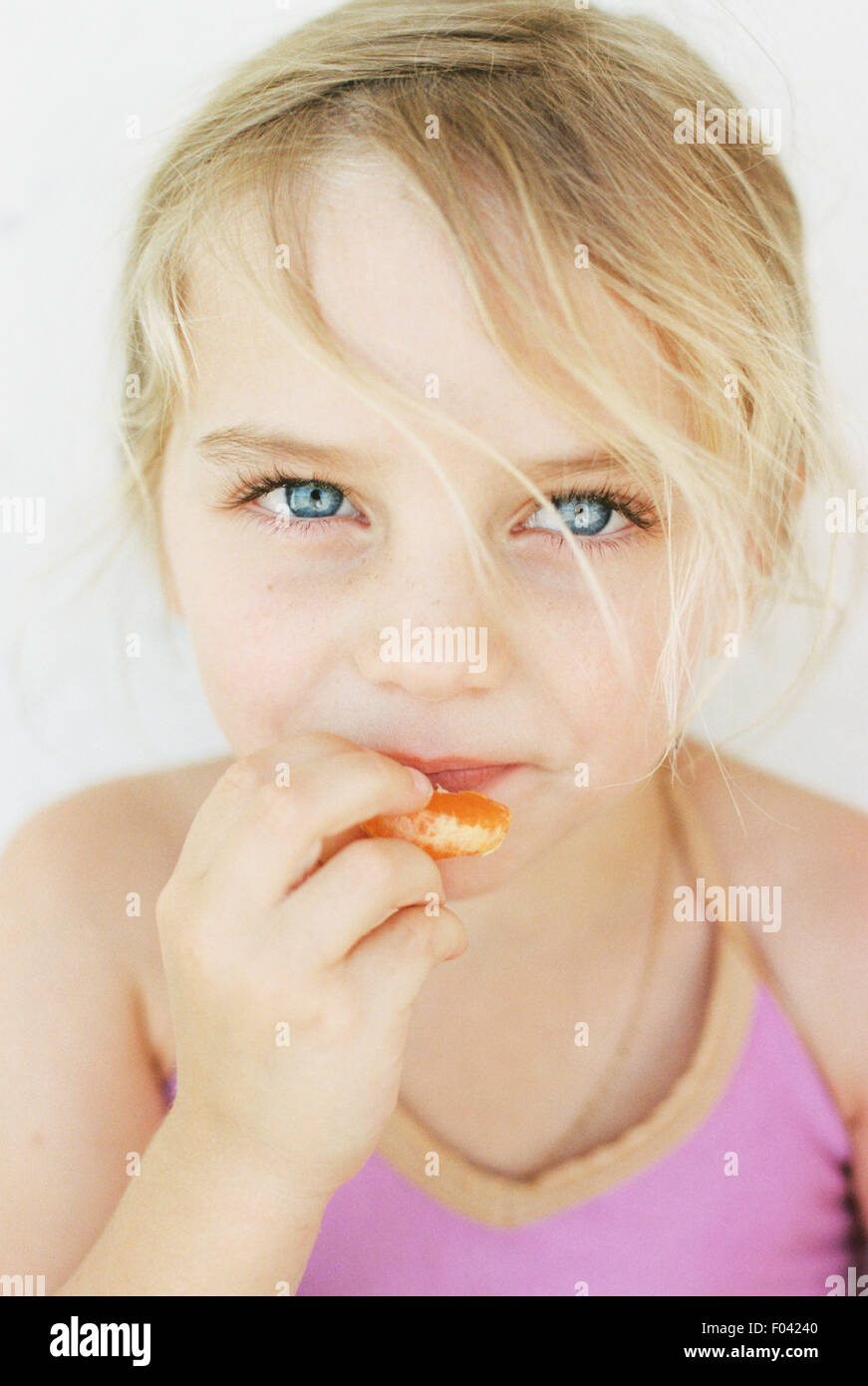 Portrait of a young blond girl, eating a mandarin, looking at the camera. Stock Photo