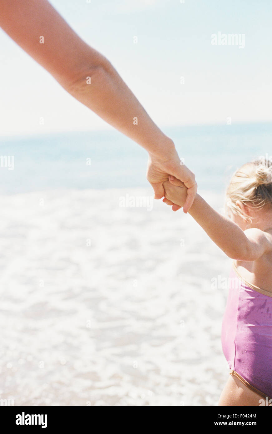 Woman and her daughter holding hands, standing on a sandy beach by he ocean. Stock Photo