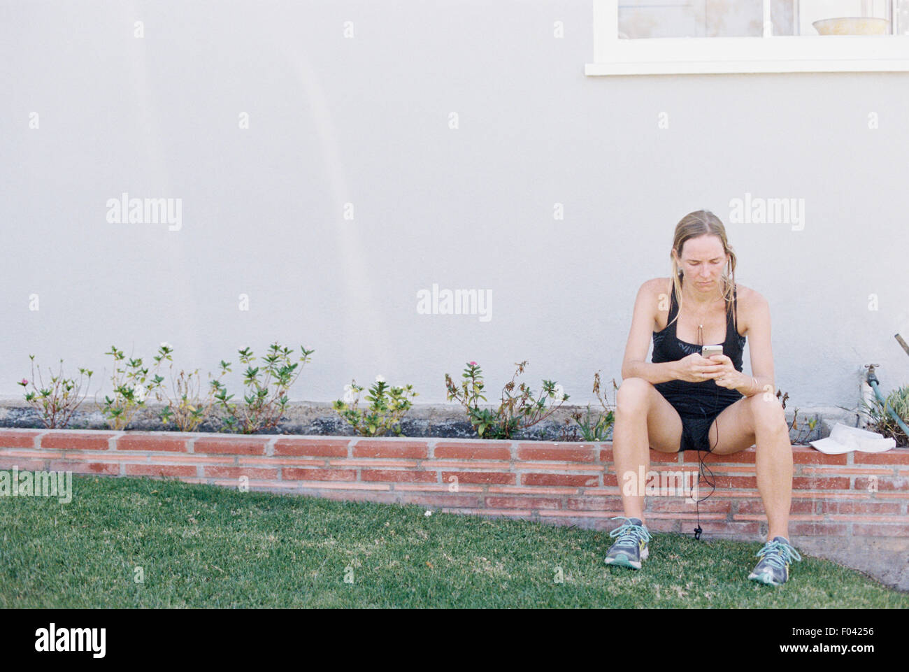 Blond woman in sportswear sitting on a garden wall, resting after a jog. Stock Photo