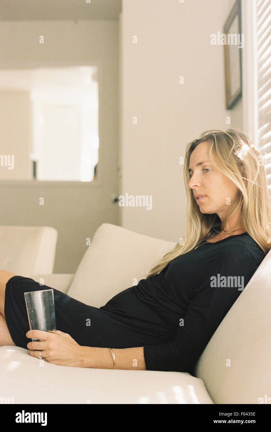 Woman with long blond hair wearing a black dress, sitting on a sofa, holding a glass. Stock Photo