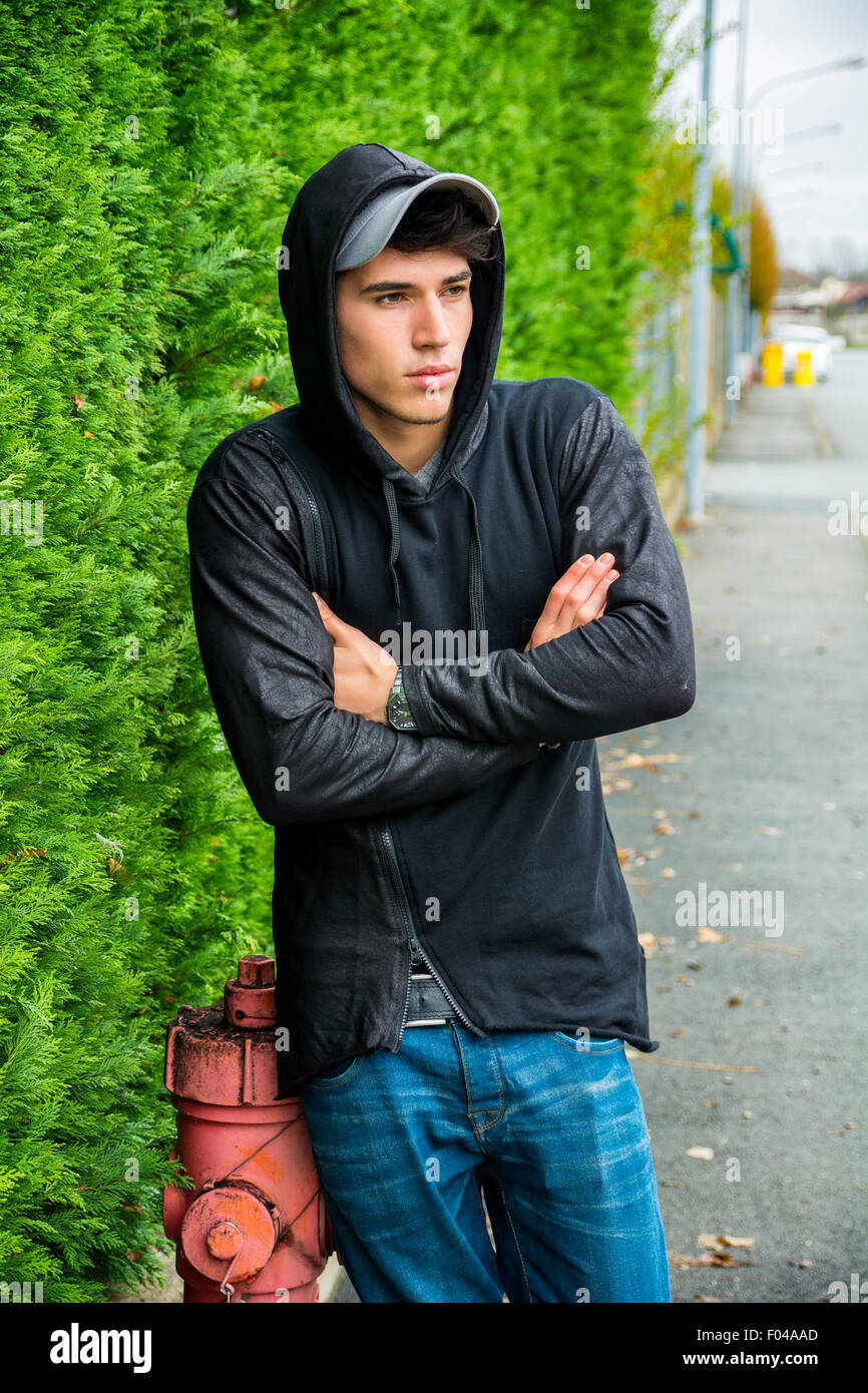 Handsome young man in black hoodie sweater standing outdoor in street looking away to a side, arms crossed on chest Stock Photo