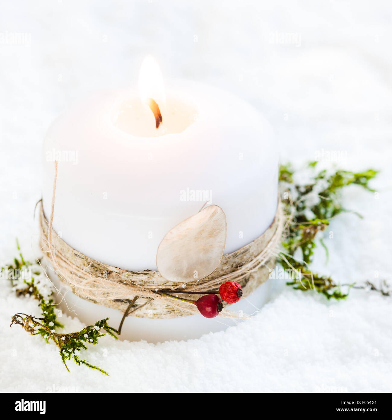 a lit white candle in the snow, decorated with birch bark, twine, red berries and honesty seedpod Stock Photo