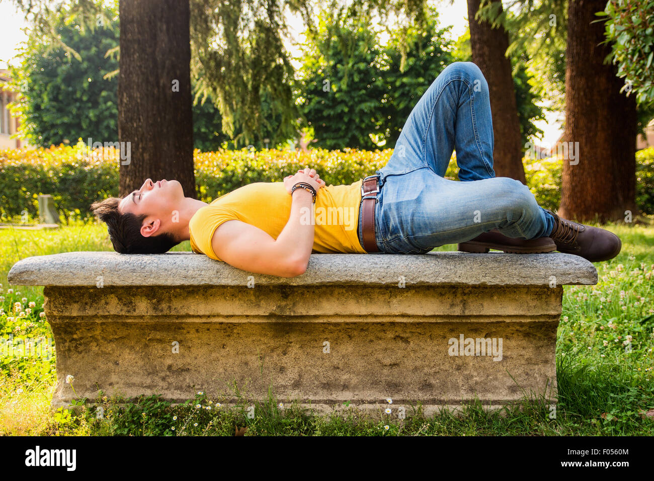 Attractive young man sleeping on stone bench outdoor in city park during day, full body shot Stock Photo