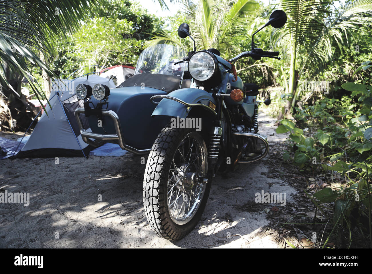 An IMZ Ural motorcycle with side car goes camping off road, on a beach amongst sand, palm trees, and tents. Stock Photo