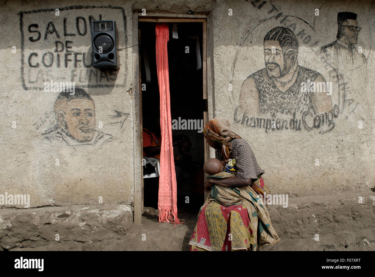 A woman with her baby sits outside a barbershop with hand painted sign in North Kivu province in DR Congo Africa. Many businesses use such paintings as a visual lingua franca to communicate what goods they sell Stock Photo