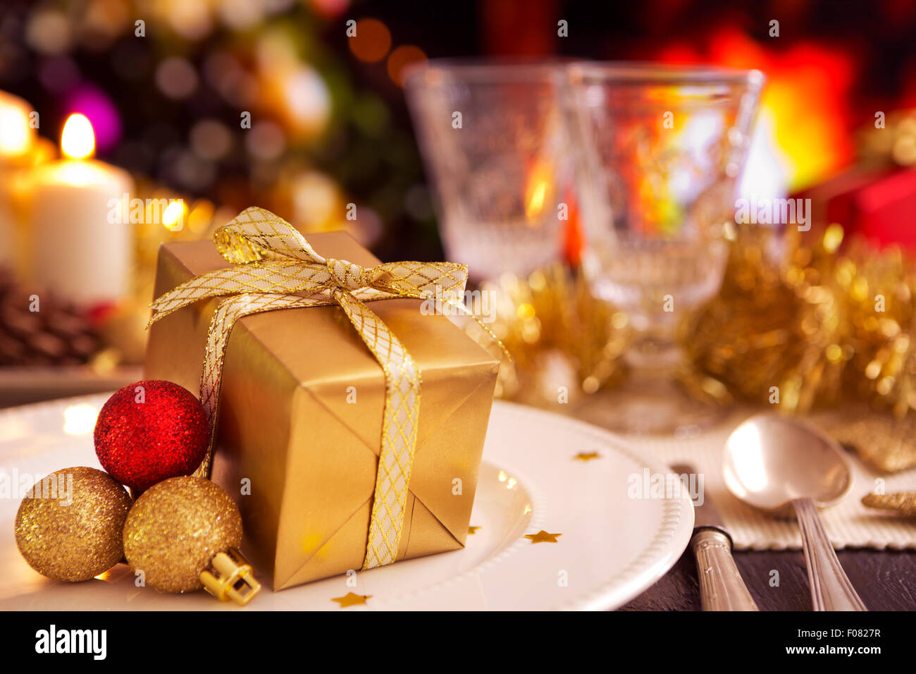 A romantic Christmas dinner table setting with a gift and Christmas decorations. A fire is burning in the fireplace in the backg Stock Photo