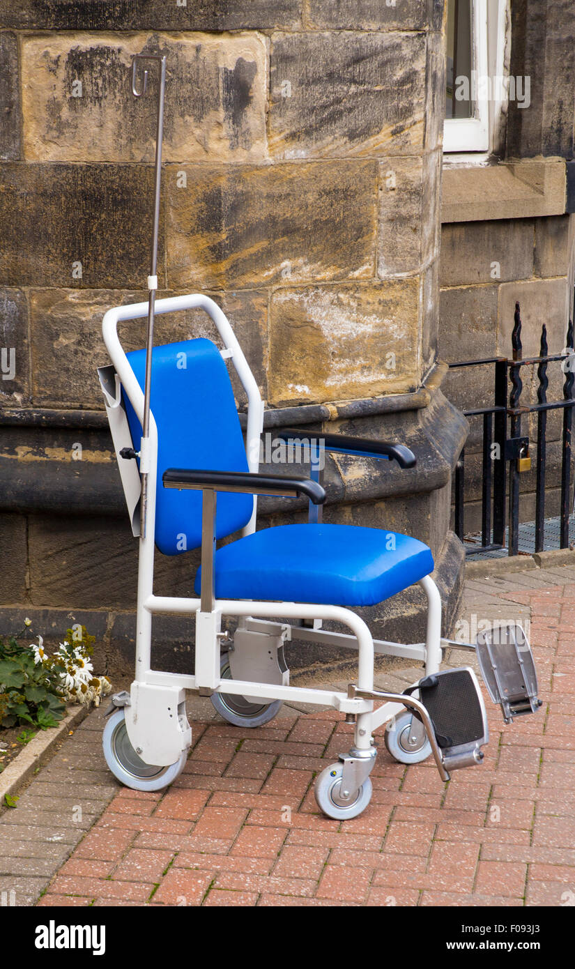 a Large Heavy Duty Wheelchair with blue seats and black arm rests and a drip stand parked outside with nobody on or in it. Stock Photo