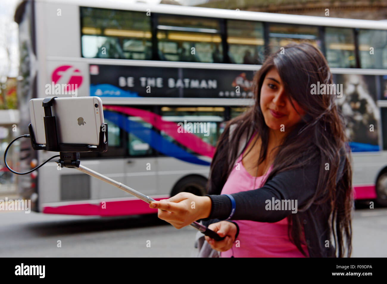 Apple iPhone held by selfie stick by out of focus young woman with double decker bus behind Stock Photo