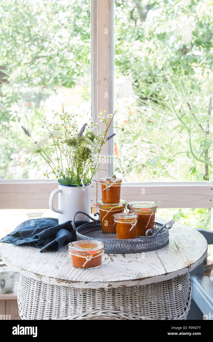 Apricot chutney jars on vintage tray, with grey kitchen towel and flower bouquet on white wooden table Stock Photo