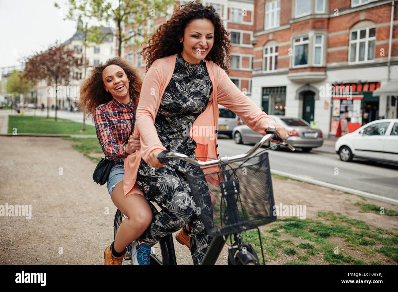 Two young women together on one bicycle having fun. Cheerful young girls enjoying cycle ride on city street. Stock Photo