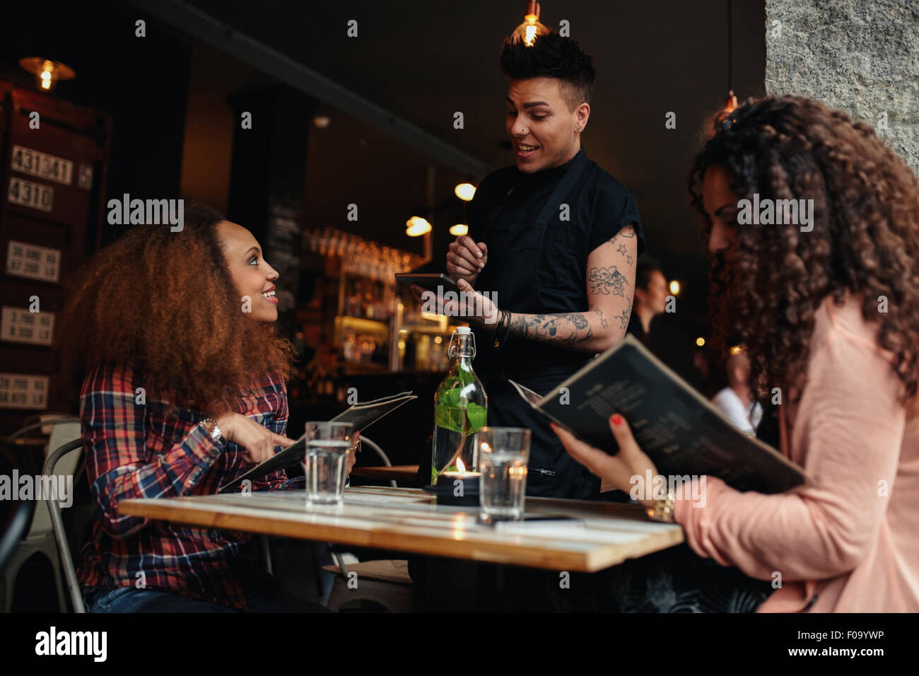 Young woman with a friend ordering to waiter holding digital tablet. Two women sitting at cafe holding menu card giving an order Stock Photo