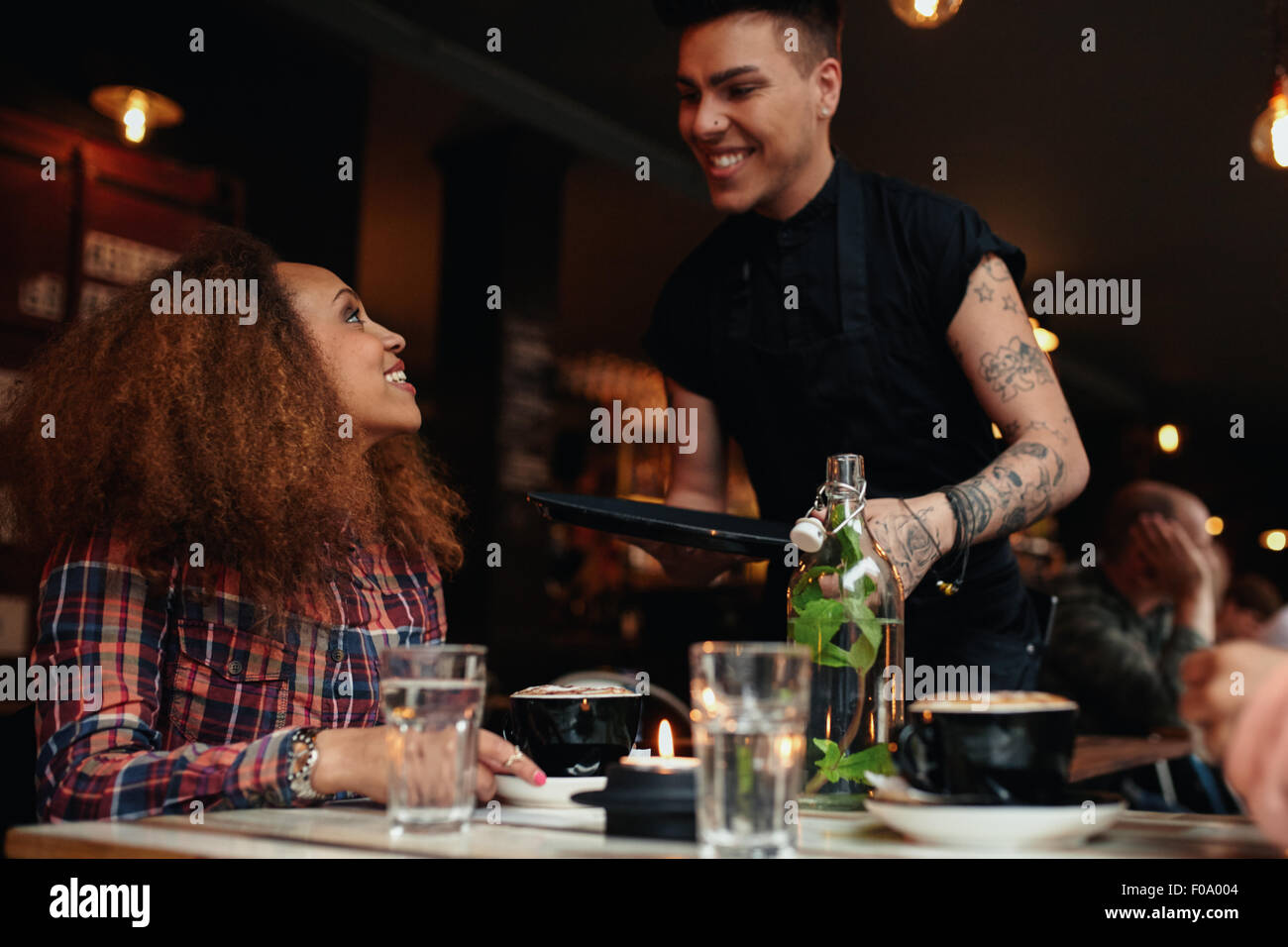 Woman talking to waiter at restaurant. Young woman sitting at cafe with waiter standing by smiling. Stock Photo