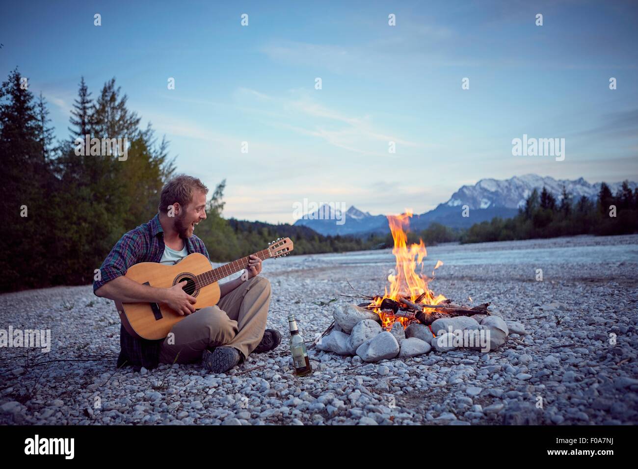 Young man sitting by campfire playing guitar, singing, Wallgau, Bavaria, Germany Stock Photo