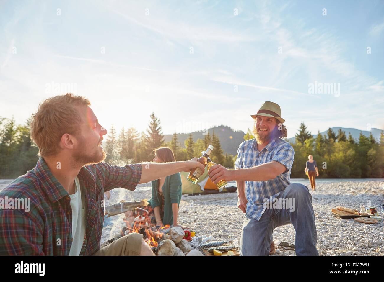 Adult men sitting around campfire making a toast with beer bottles Stock Photo