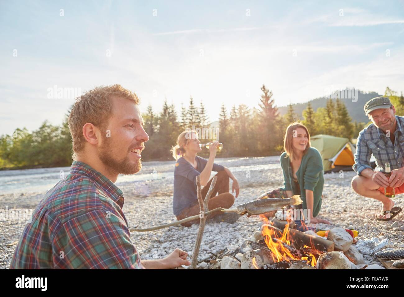 Adults sitting around campfire drinking bottles of beer, smiling Stock Photo