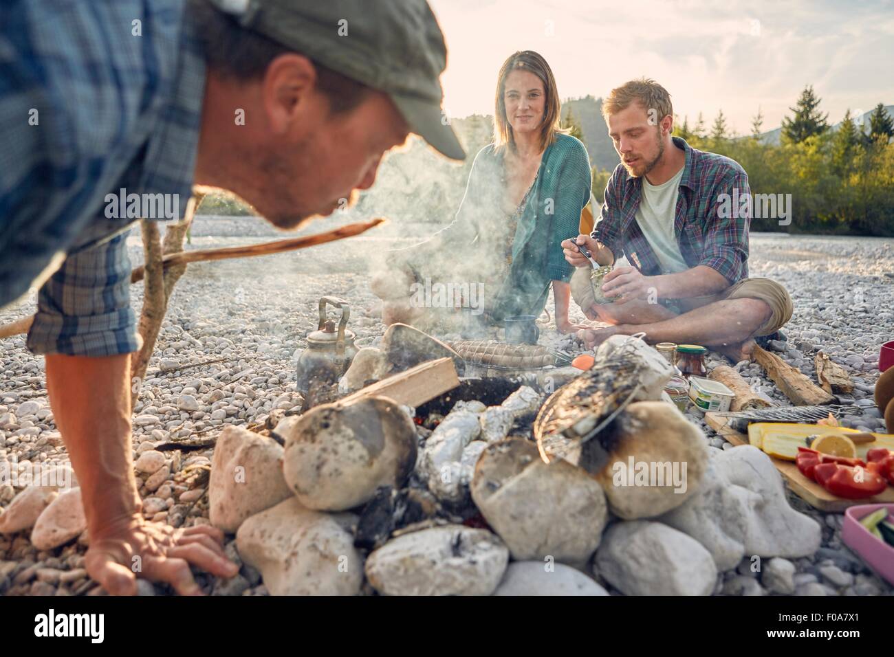 Adults sitting around campfire preparing food and blowing on embers Stock Photo