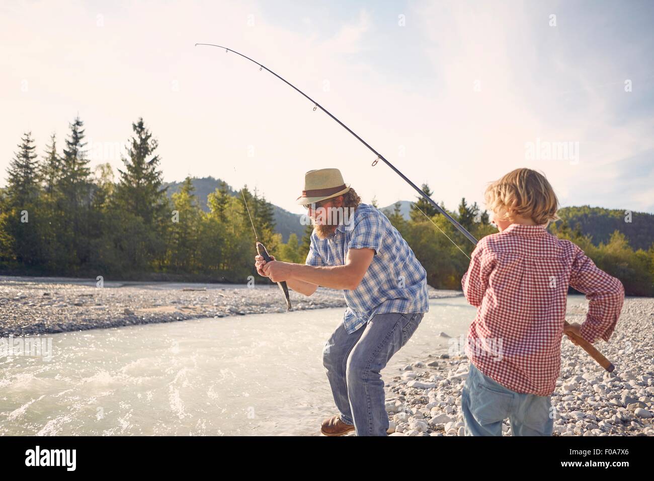 Mid adult man and boy next to river using fishing rod to catch fish Stock Photo
