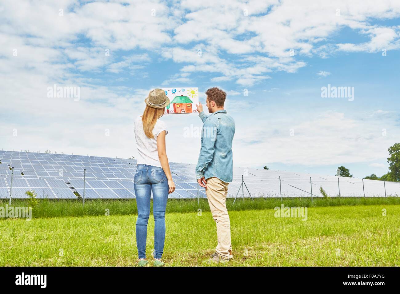 Young couple standing in field, looking at child's drawing of house, next to solar farm Stock Photo