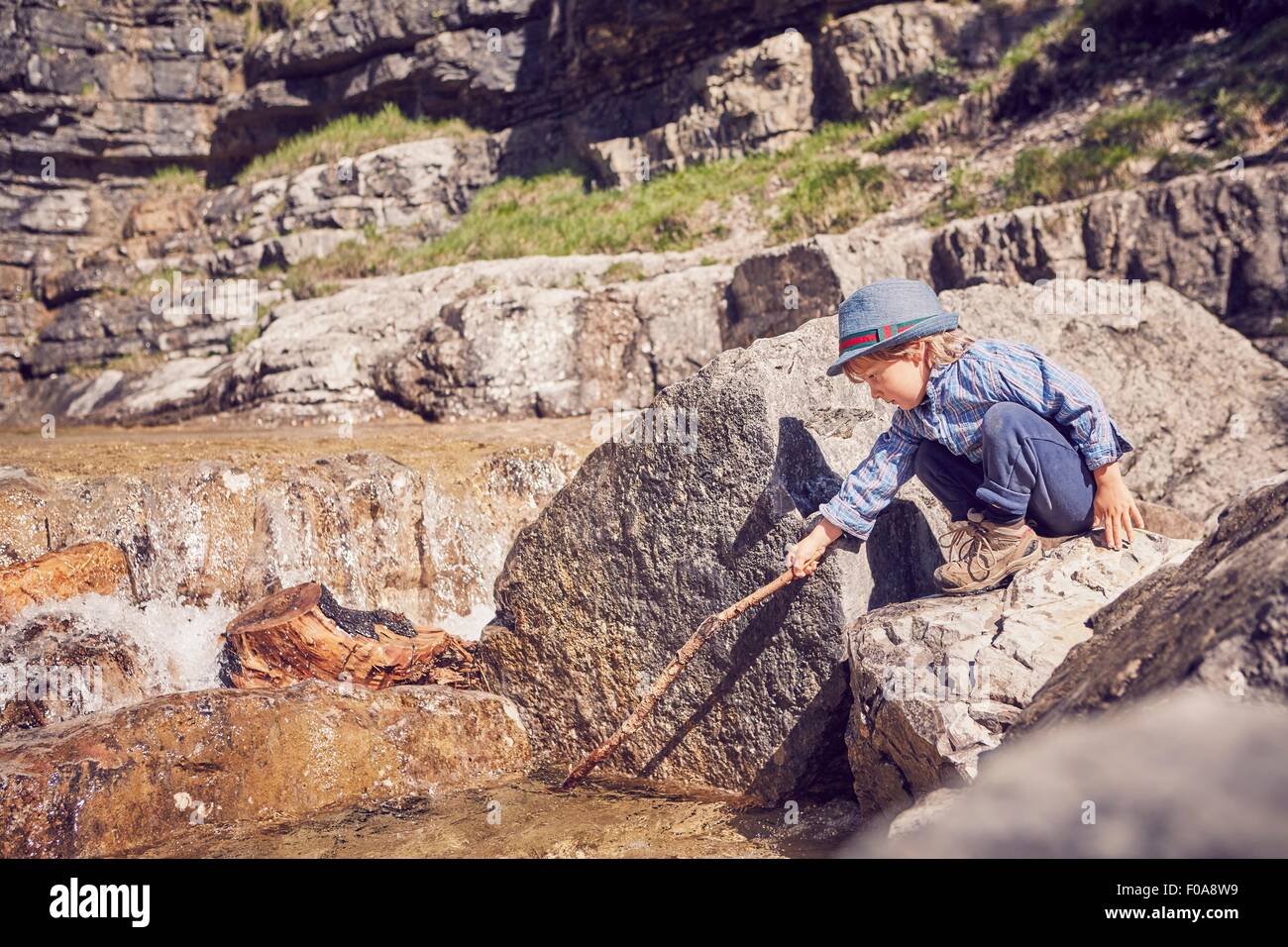 Young boy, sitting on rock, prodding mud with stick Stock Photo