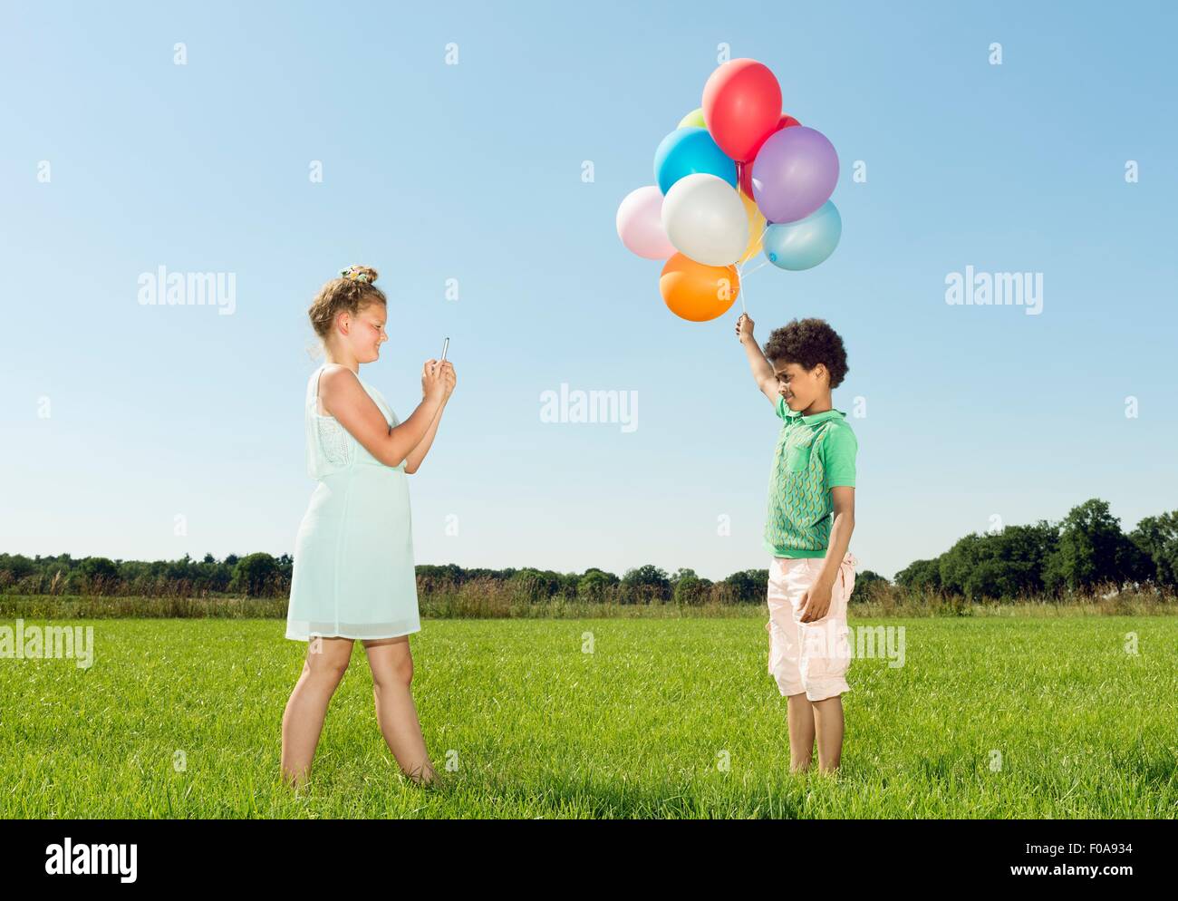 Girl taking smartphone photograph of boy holding bunch of balloons in field Stock Photo