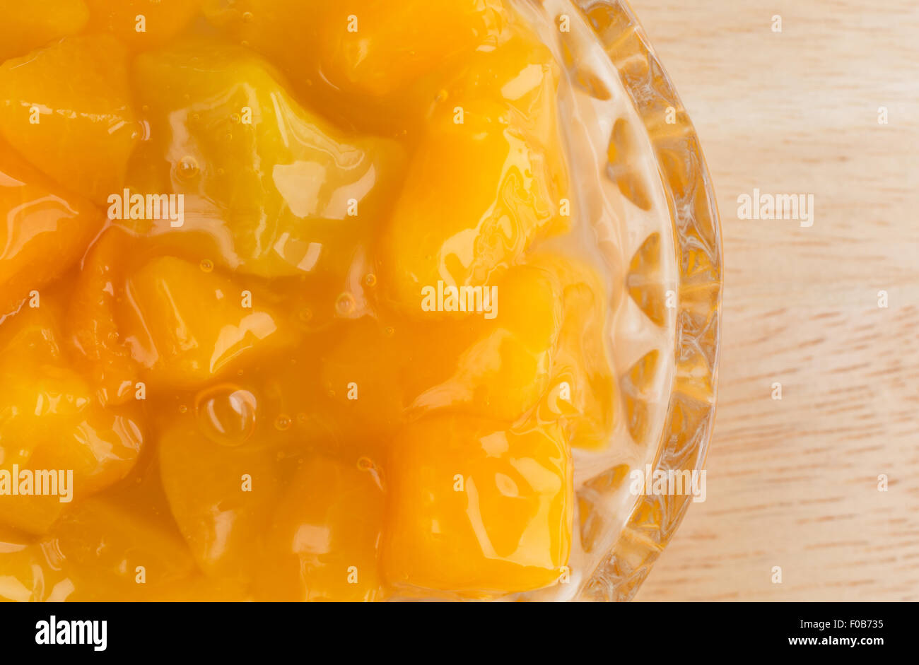 Top close view of a small glass bowl of diced peaches in heavy syrup on a wood table top illuminated with natural light. Stock Photo