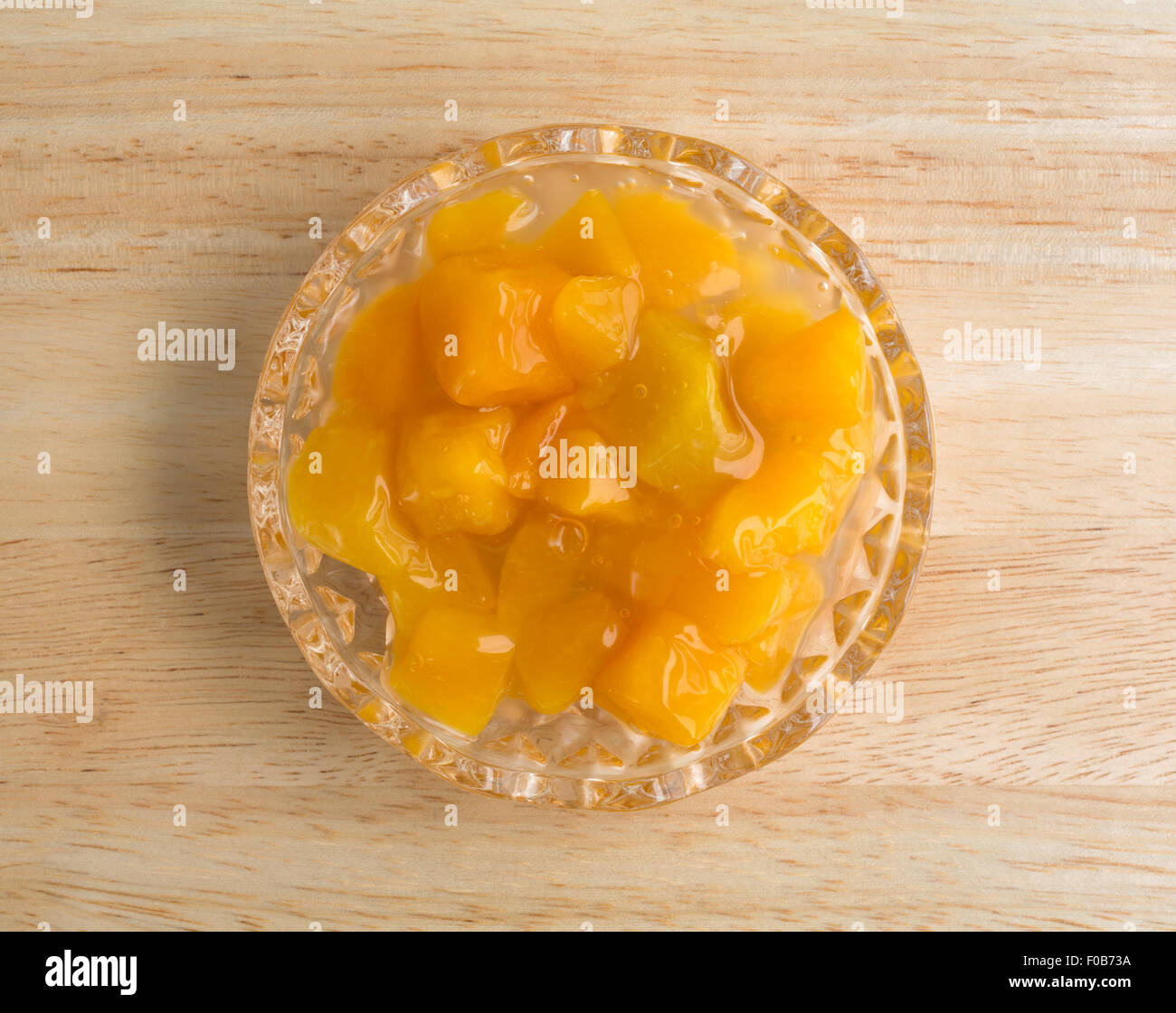 Top view of a small glass bowl of diced peaches in heavy syrup on a wood table top illuminated with natural light. Stock Photo