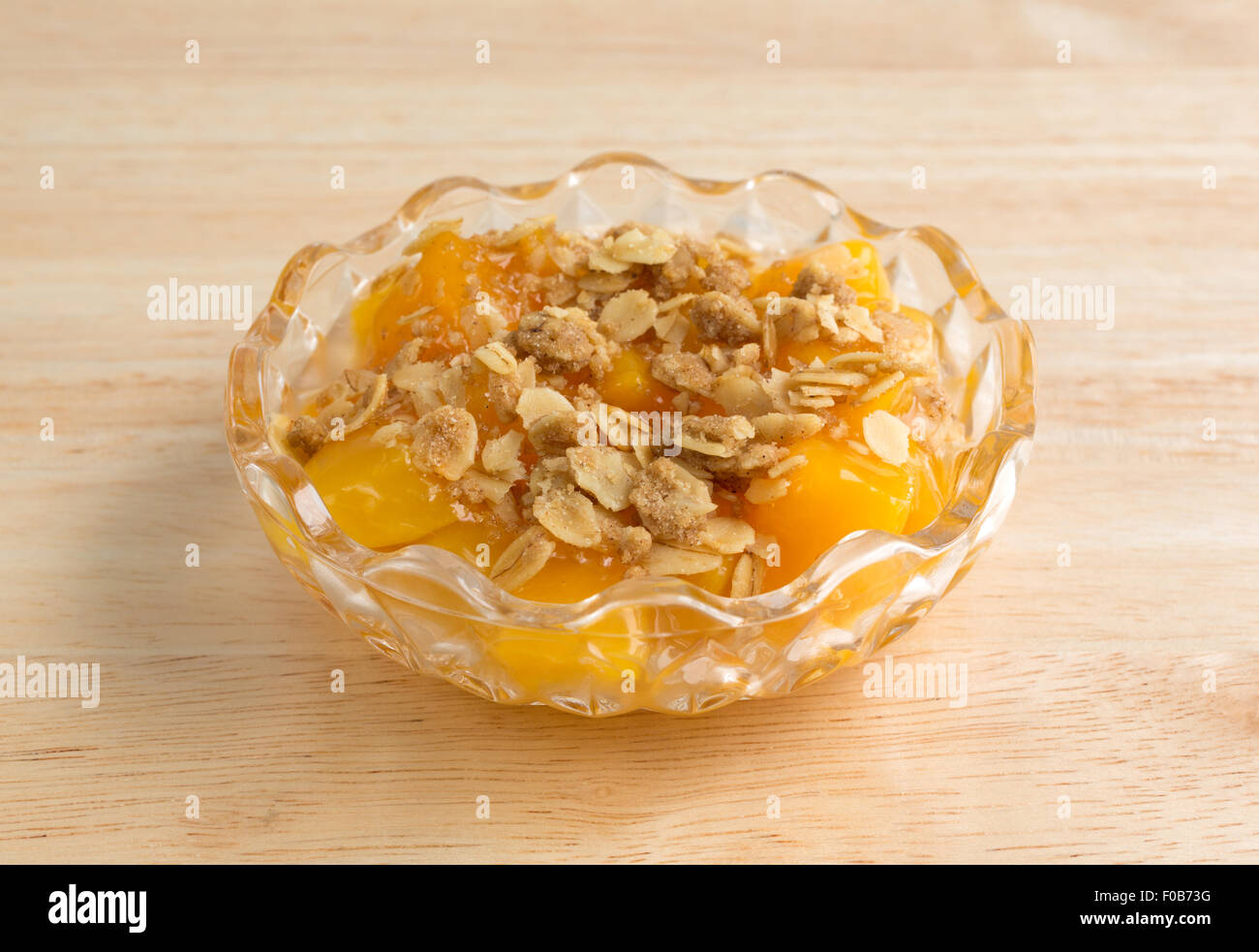 A small glass bowl of diced peaches in heavy syrup with oats and brown sugar topping on a wood table top Stock Photo