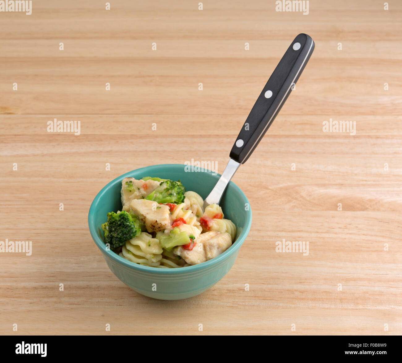 A small bowl with a serving of grilled chicken chunks and assorted vegetable in a garlic sauce with a fork inserted atop a wood Stock Photo