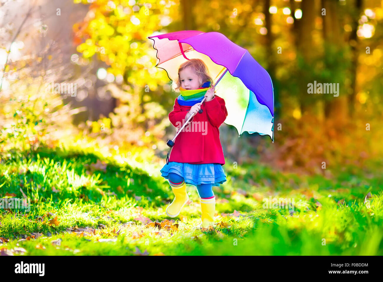 Little girl playing in the rain in autumn park. Child holding umbrella walking in the forest on a sunny fall day Stock Photo