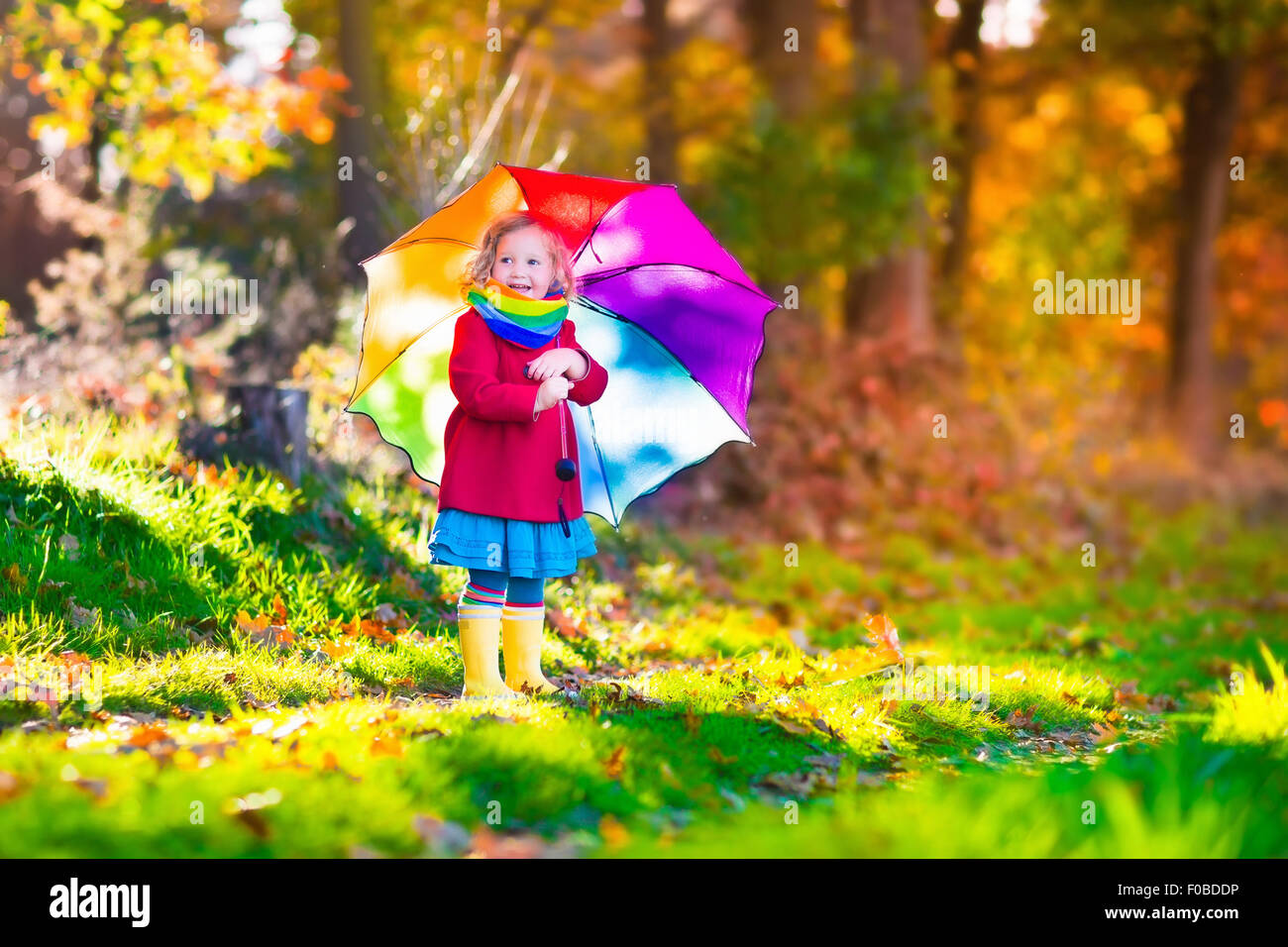 Little girl playing in the rain in autumn park. Child holding umbrella walking in the forest on a sunny fall day Stock Photo