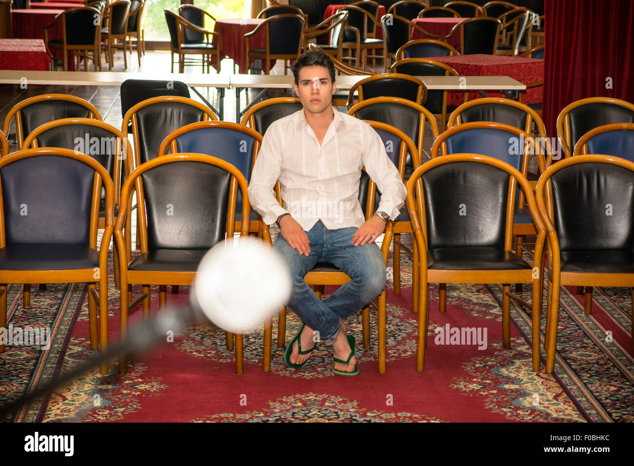 Handsome Young Man in Casual Wear, Sitting at the Audience Chair In Front of a Podium Alone and Looking at the Camera. Stock Photo