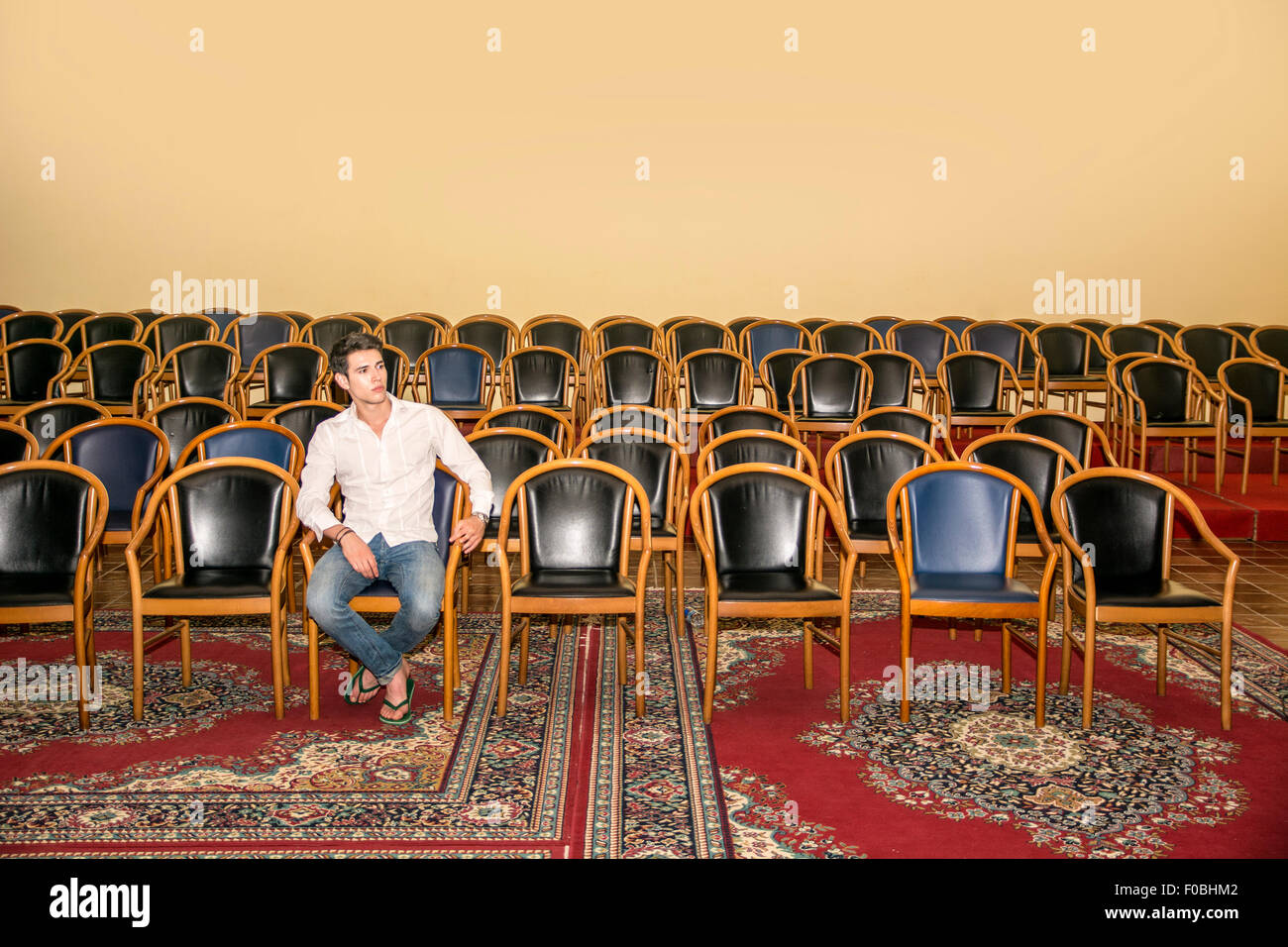 Handsome Young Man in Casual Wear, Sitting at the Audience Chair In Front of a Podium Alone and Looking at the Camera. Stock Photo