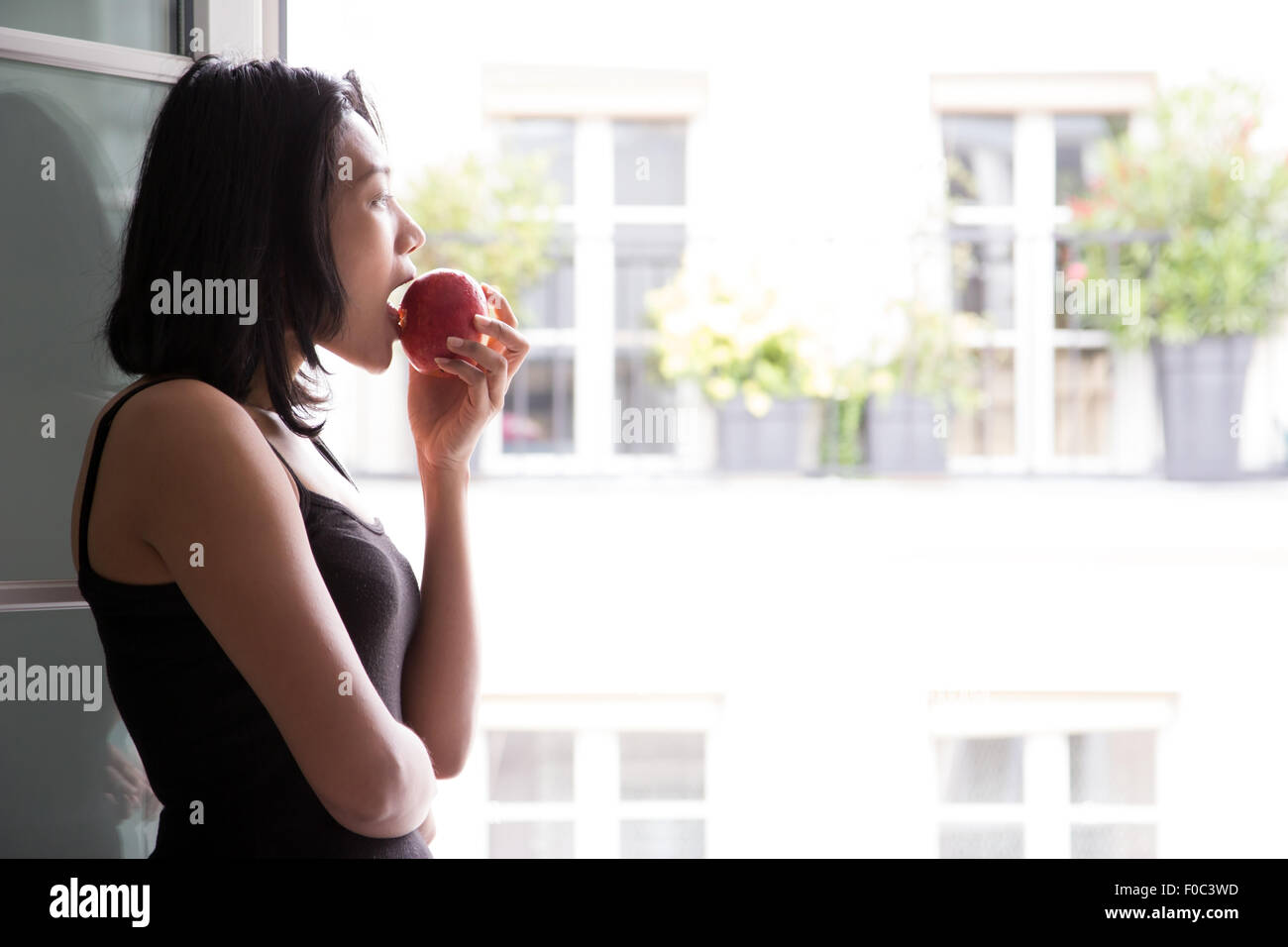 Young woman standing at an open window and eat apple Stock Photo