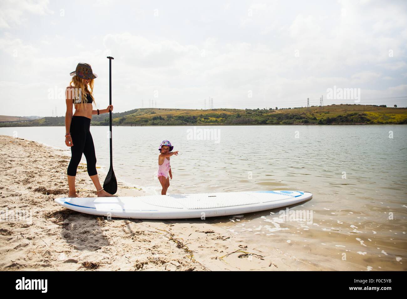 Mid adult woman and toddler daughter with paddleboard on beach, Carlsbad, California, USA Stock Photo