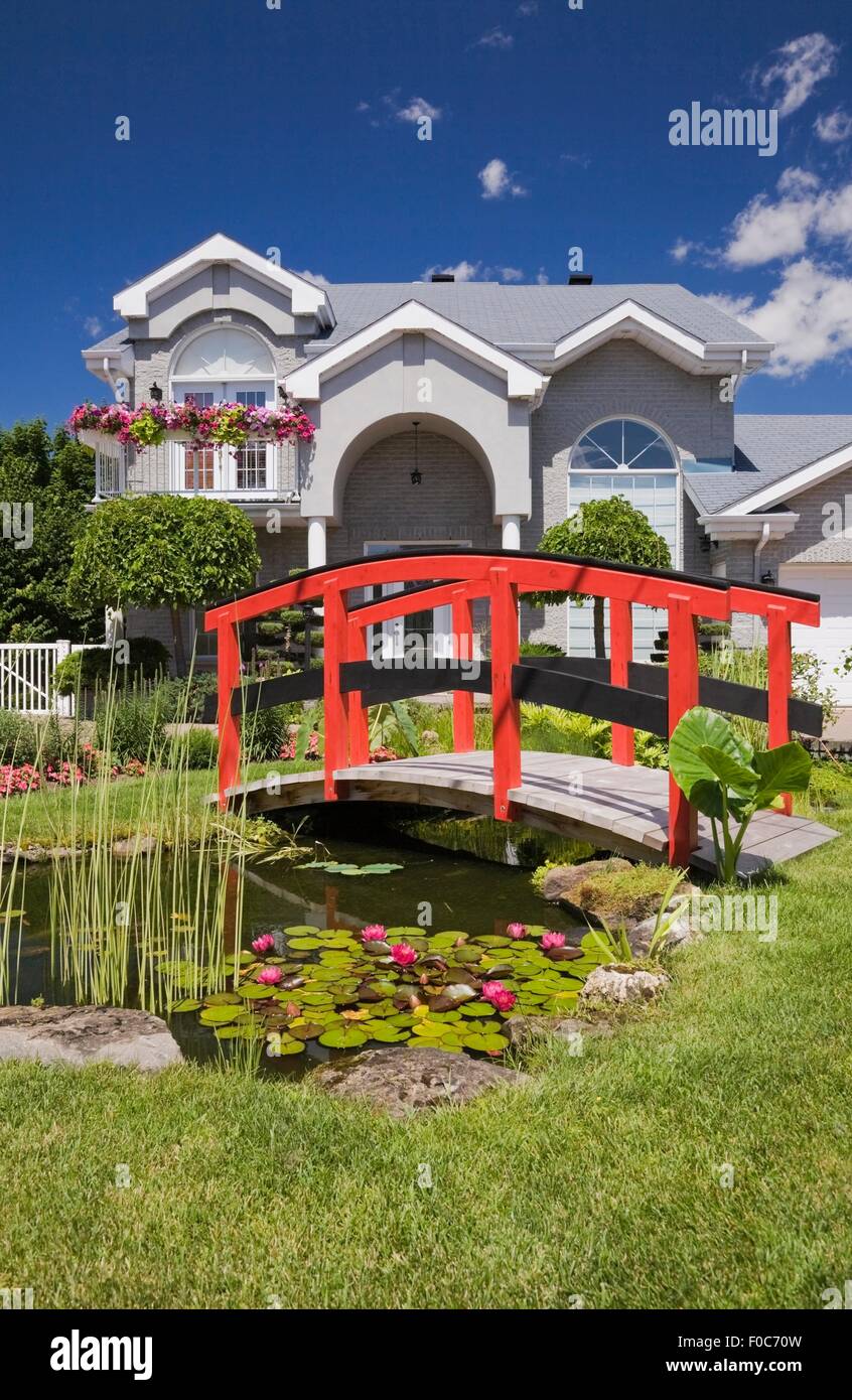 Grey brick cottage style residential home and landscaped  manicured garden with red wooden footbridge, Quebec, Canada Stock Photo