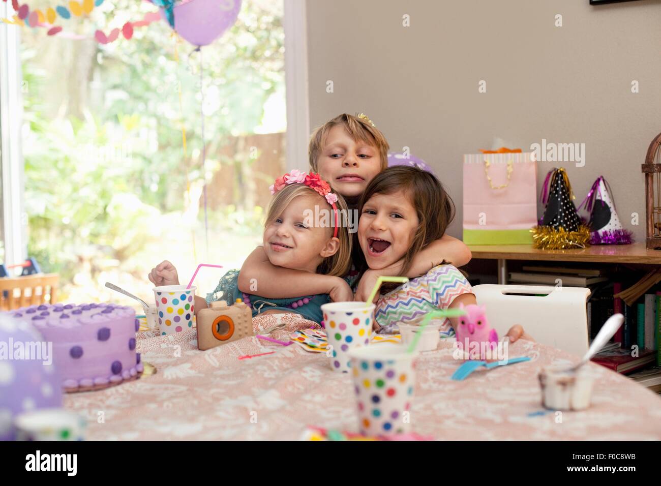 Three children sitting at birthday party table hugging each other Stock Photo