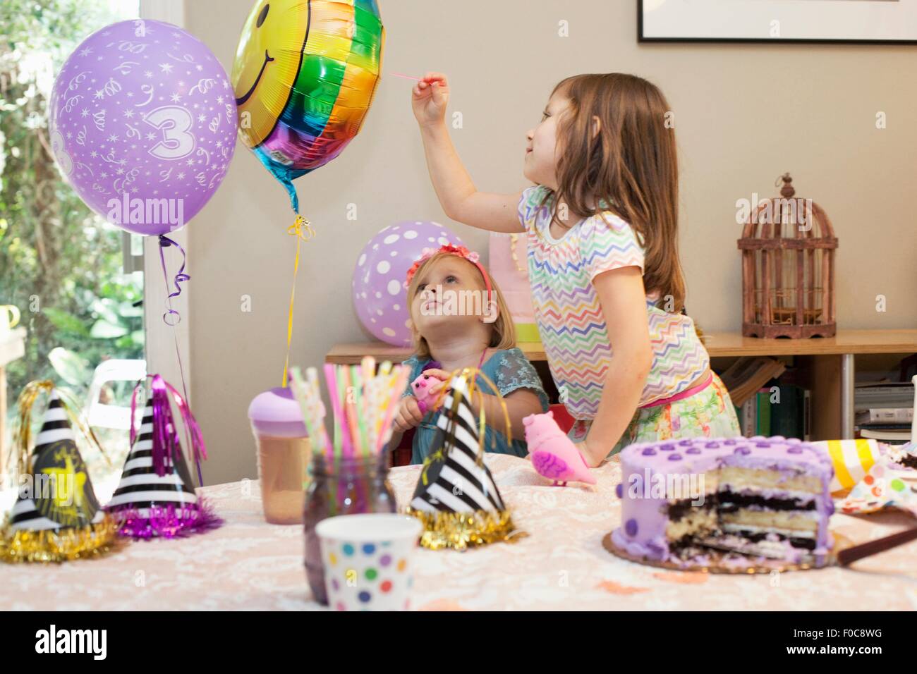 Two girls sitting at birthday party table with cake playing with balloons Stock Photo