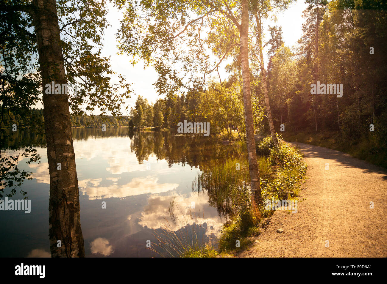 Warm temperature landscape, gravel road next to perfectly still lake. Reflections of clouds in the water. Stock Photo