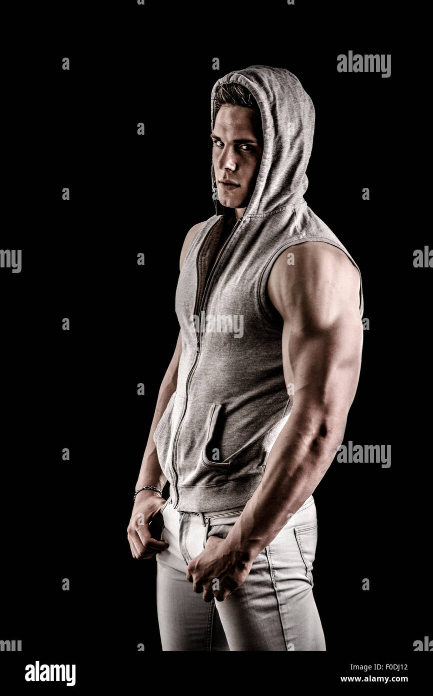 Handsome muscular tough young man in dark hooded t-shirt isolated on black background, looking at camera Stock Photo