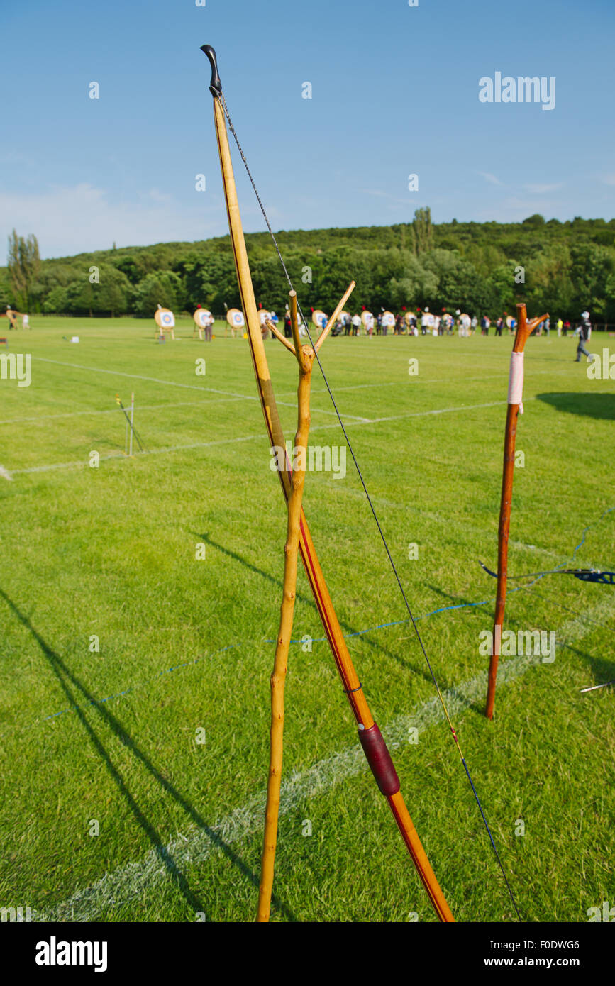 Traditional wooden longbow resting on stand during competition event with archers retrieving arrows from targets other side of f Stock Photo