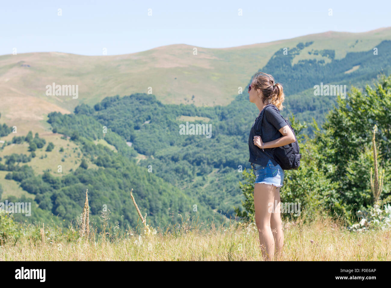 Young, beautiful girl with a backpack on her back, standing on the plateau. Green meadows and majestic mountains in the backgrou Stock Photo
