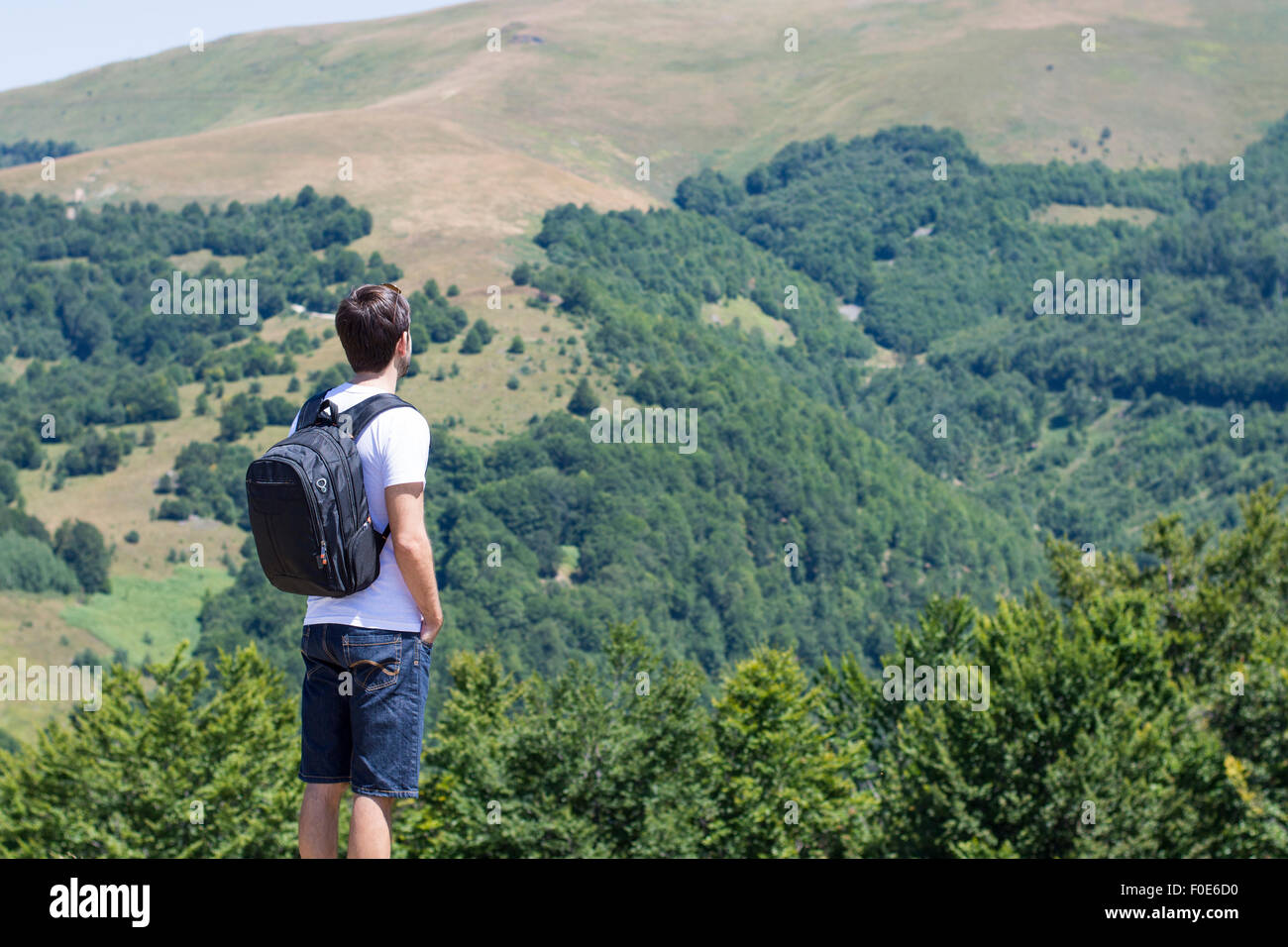 Young man with backpack standing on cliff's edge and looking to the mountains. Hiking adventure Stock Photo