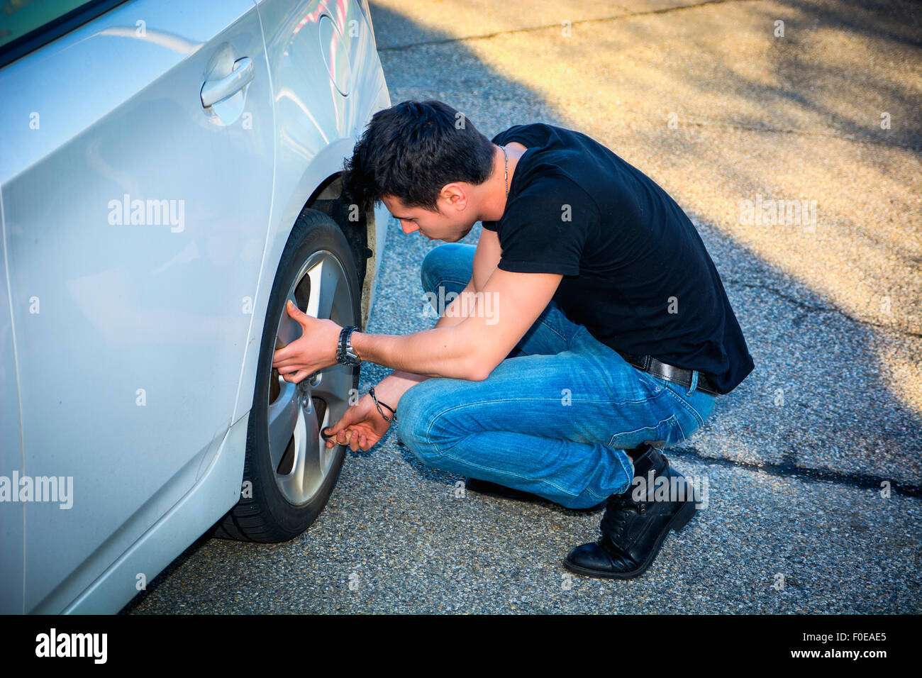 Handsome Young Man in Casual Clothing Squatting Besides the Wheel of a his White Car Changing Tires Stock Photo